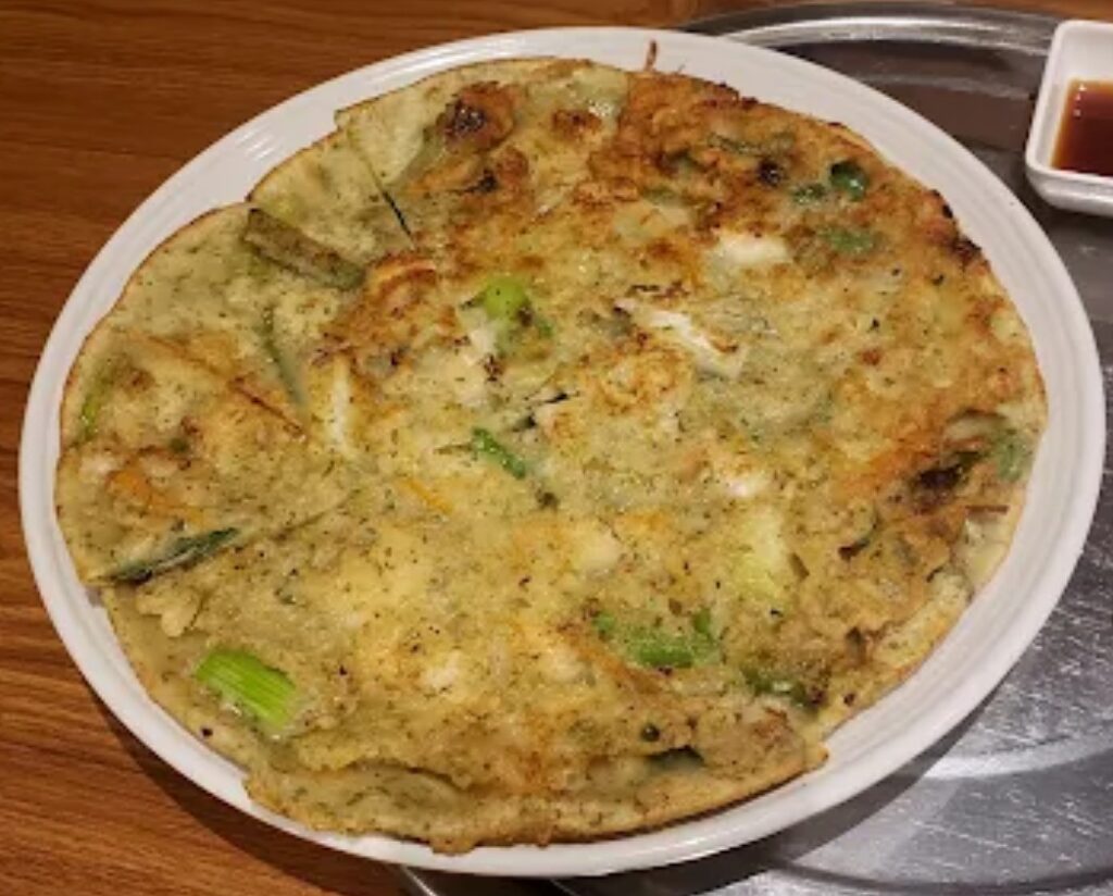 A golden-brown Korean seafood pancake from Alirang, served on a dark plate; thinner than expected with minimal crispiness and visible seafood ingredients.