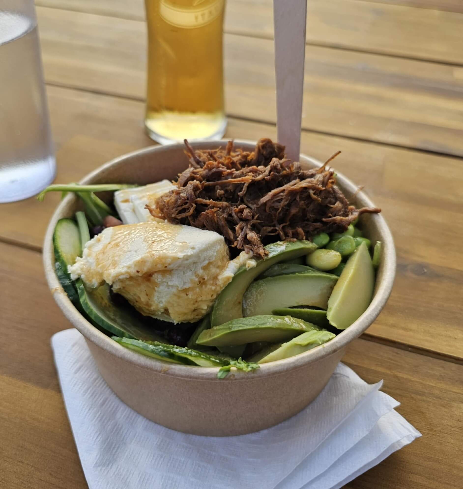 A compostable bowl filled with pulled beef, avocado, cucumber, edamame, and greens, placed on a napkin on a wooden table with a glass of water and a beer in the background.