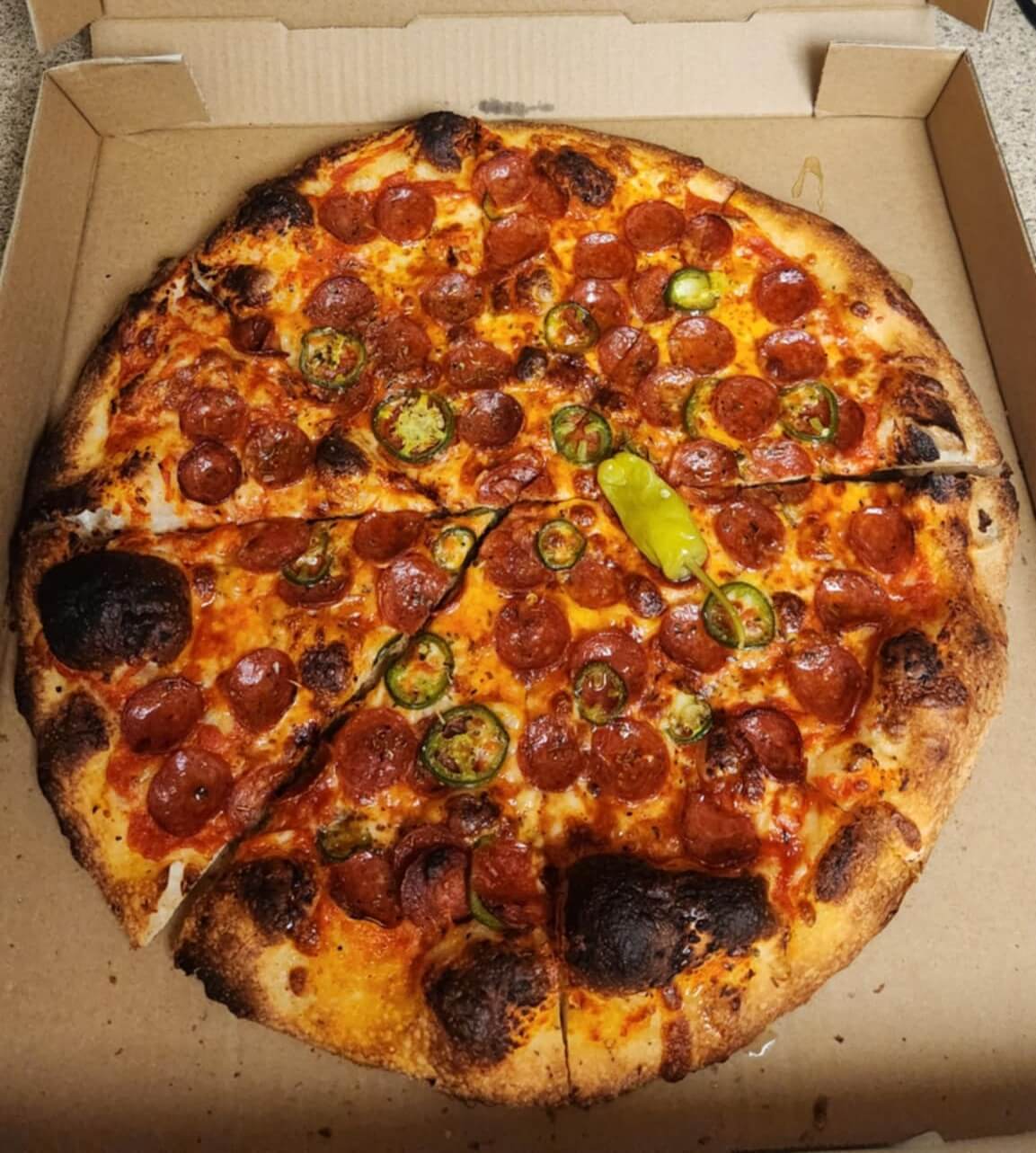 A large Sweetheart pizza from Heartbreakers Pizza in an open box, topped with pepperoni, jalapeño slices, and a whole pepper, featuring a charred, crispy crust and a golden cheese finish.