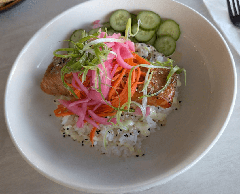 A miso-glazed salmon bowl served with rice, sesame seeds, pickled onions, shredded carrots, green onions, and sliced cucumber at Seafood Grill.