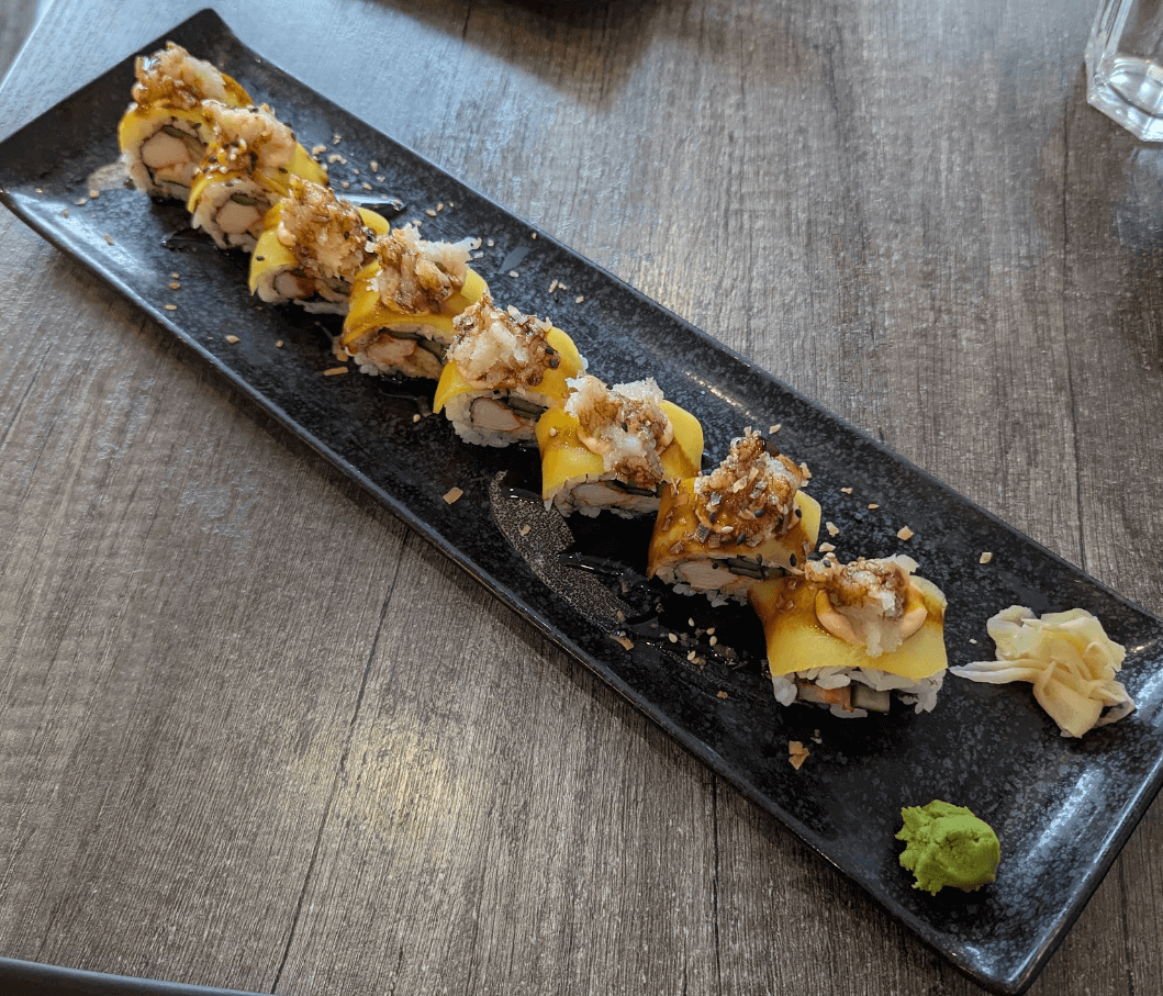 Tropicana Roll sushi topped with mango slices, imitation crab, and crunchy bits, served with wasabi and pickled ginger on a black ceramic plate at Sushi-ro