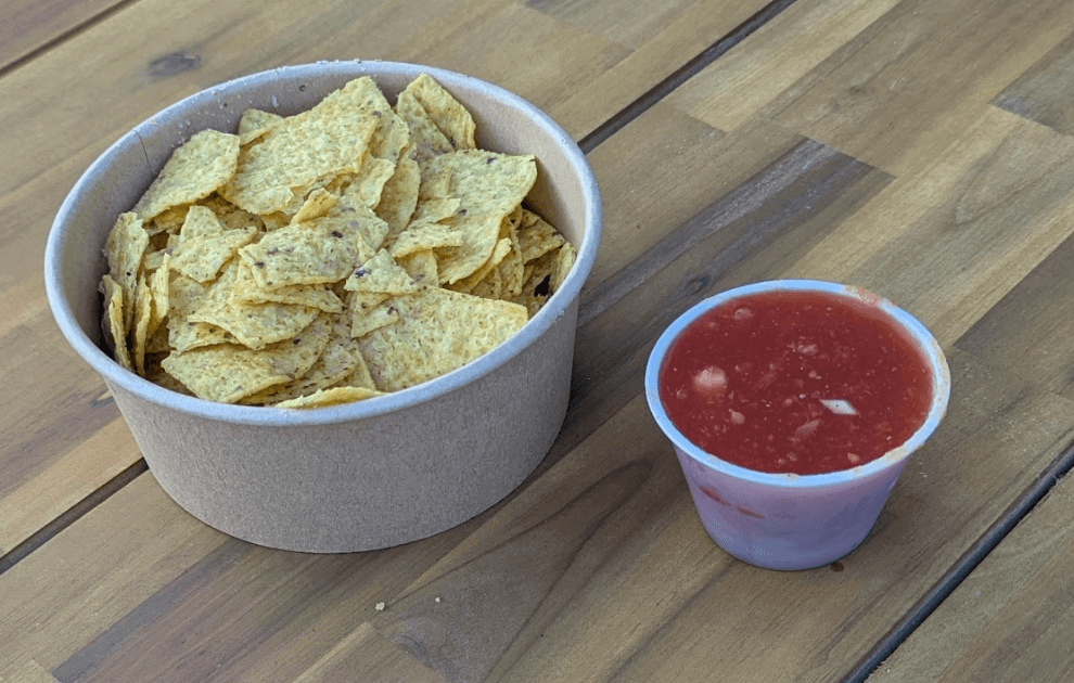 A brown paper container filled with tortilla chips next to a small plastic cup of red salsa on a wooden table.